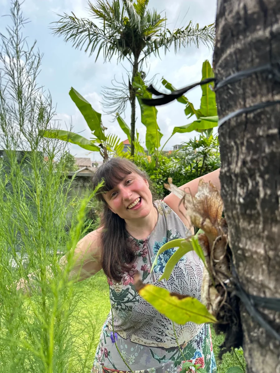 Nadja lächelt zwischen tropischen Pflanzen neben einem Baum im Garten